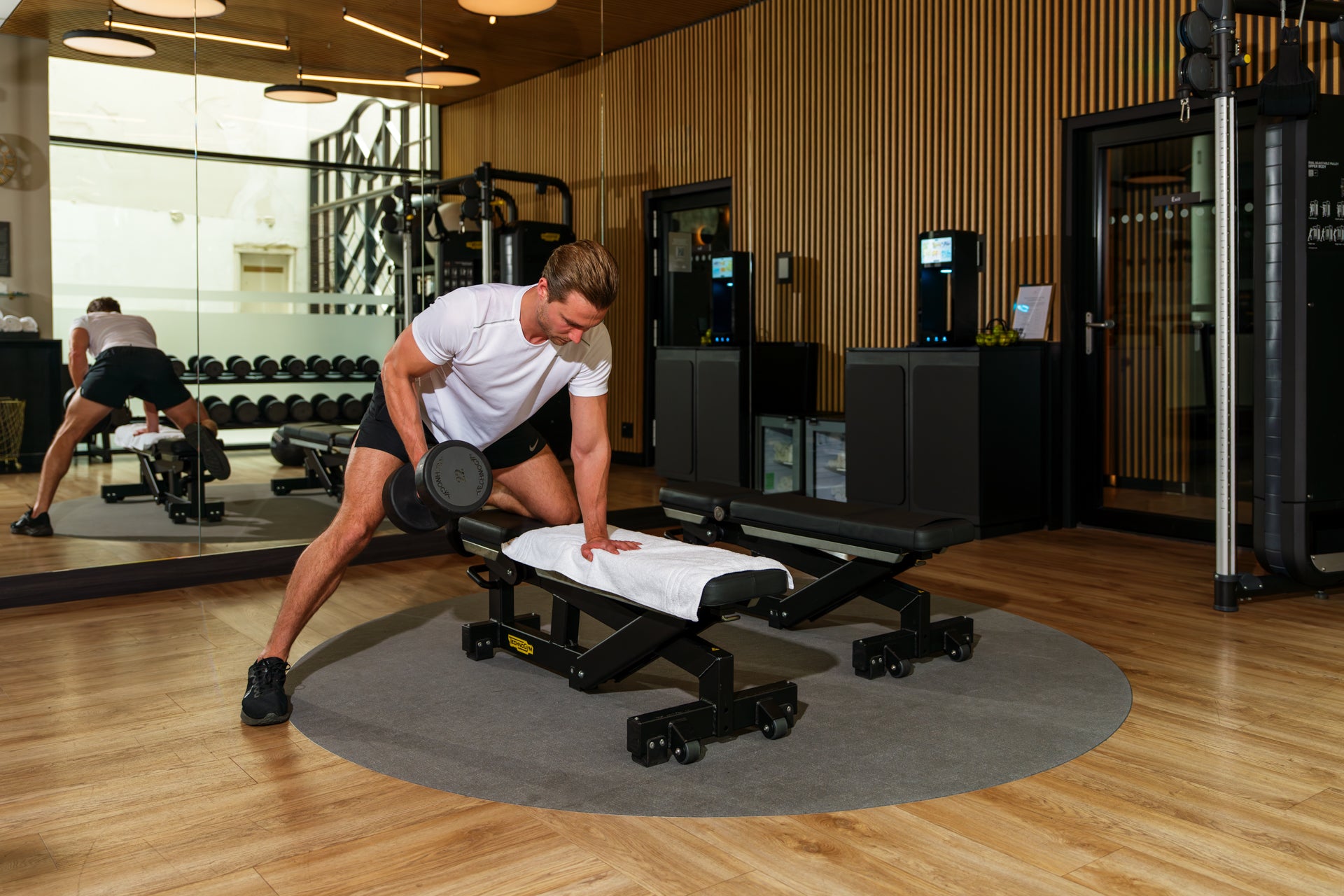 Man voert een roeibeweging met dumbbell uit terwijl hij op een halterbank steunt, met de spiegelwand achter hem